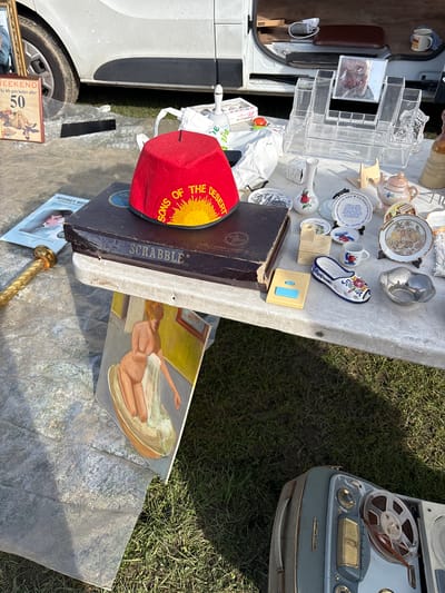 This image depicts a collection of items displayed on a table, likely at a flea market or yard sale. The location is near Chirk, United Kingdom. Items include a Scrabble game, a fez hat with "Sons of the Desert" text, an old tape recorder, decorative plates and mugs, an acrylic organizer, a painting of a nude figure, and other assorted bric-a-brac. The table is set up near a vehicle, and the surrounding environment includes grass and a paved surface. There are no people visible in the image.