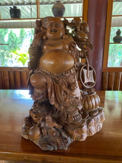 The image shows a large, intricately carved wooden statue of a jovial, rotund figure, likely representing the Budai or "Laughing Buddha". The statue is predominantly made of polished, light brown wood and is positioned on a wooden table. The Budai figure is depicted with a wide smile, a prominent belly, and is adorned with flowing robes. Around him are various carved objects including gourds, fruits, and smaller figures of children. One hand holds a Ruyi scepter, a ceremonial object, while another appears to be resting on a large sack. The carving is detailed, with textures and patterns visible on the robes and surrounding elements.
The setting appears to be indoors, with hints of natural light coming from windows. Through the windows, lush green foliage and trees are visible, suggesting an outdoor environment with abundant greenery. The interior elements include wooden paneling and some decorative ceramic gourds hanging from the ceiling, visible through the window. The lighting suggests it is daytime, with soft, natural light illuminating the statue. The overall impression is one of peace, prosperity, and good fortune, often associated with the Budai figure. The scene is static, with the statue being the sole subject of focus. No visible text is present in the image.