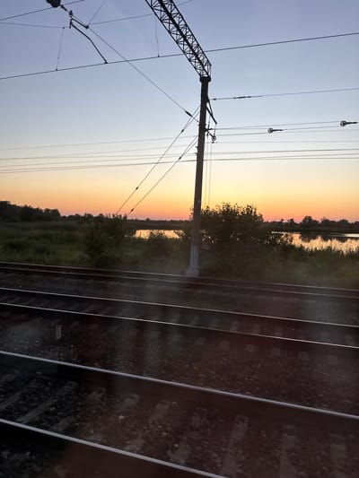 The image shows a train track in a rural landscape during sunset. The sky is a gradient of pale blue to orange, with the sun low on the horizon. There are power lines and a utility pole visible above the train tracks. To the right of the tracks, there is a body of water and a dense line of trees and vegetation. The train tracks themselves are the foreground, with ballast and sleepers clearly visible. The scene is likely located in a rural area near Enem, Russia, given the context. There are no people or animals visible in the image.