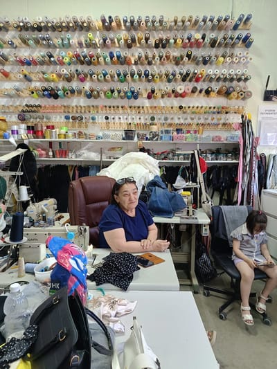 In Novaya Adygeya, Russia, a sewing workshop is depicted. A middle-aged woman, wearing a blue t-shirt and sunglasses on her head, sits at a sewing machine. She appears to be engaged in work or overseeing activities. To her right, a young girl, dressed in a striped shirt and shorts, sits on a chair and looks at a mobile phone. The workshop is filled with spools of thread mounted on the wall, fabric, sewing machines, and other sewing supplies. The overall impression is of a functional workspace dedicated to tailoring or garment creation.