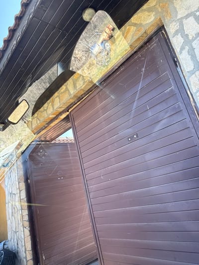 The image displays an exterior scene in Bezden, Bulgaria, viewed from inside a vehicle through its windshield. The primary subject is a two-panel, dark brown metal gate system with horizontal slats, partially open. The right gate panel is largely closed, featuring a small, rectangular black control unit with two light-colored circular elements near its center. The left gate panel is ajar, revealing a passage and a glimpse of a terracotta-tiled roof structure within. Above the gates, a building facade constructed of irregular, light-toned stone blocks is visible, topped by a dark wooden soffit with terracotta roof tiles. A circular, light-colored device, possibly a security camera or light fixture, is mounted beneath the wooden soffit.

The vehicle's windshield reflects internal elements and the external environment. A side-view mirror is reflected on the left, showing a distant natural landscape under a blue sky. Prominent reflections on the windshield include a white object with red accents, resembling a small toy vehicle, in the upper center, and a rectangular label or sticker with the text "BG 03 BA 9369" in the lower left. Sunlight creates glare and streaks across the windshield. To the far left, outside the vehicle, a stack of vehicle tires is partially visible against a stone wall. No individuals are depicted.