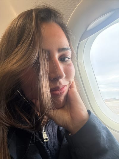 A woman is posing for a selfie in the window seat of an airplane. The window frame and part of the exterior can be seen above her. The woman is light-skinned with dark eyes and brown hair. She is wearing a dark blue "POLO" jacket. The location is not specified but it's stated that the city is San Antonio in the United States of America.
