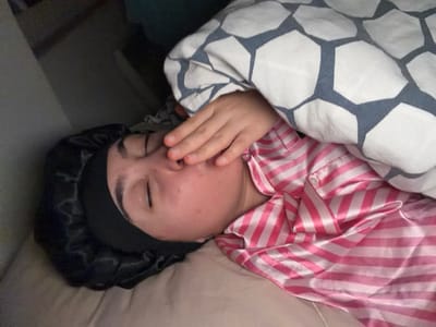 A person lies in bed, partially covered by a blanket with a white and grey geometric pattern. They wear a black satin bonnet and a pink and white striped satin pajama top. Their eyes are closed, and their hand is near their face. The lighting suggests it might be morning or a dimly lit room.