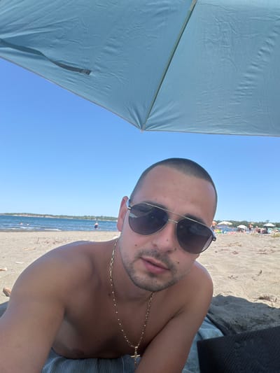 A young man with a buzz cut and sunglasses lies on a beach under a light blue umbrella. He is wearing a gold chain with a cross pendant. The beach is sandy, with some debris scattered on it. In the background, the sea is visible, and several beach umbrellas and people are seen further down the shoreline. The sky is clear and blue. The location is identified as Unknown, Bulgaria.