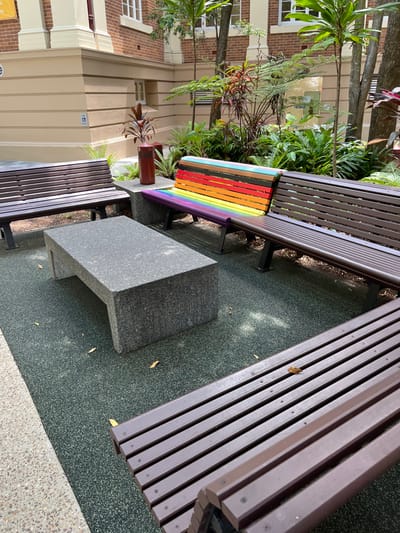 This image, taken in Brisbane City, Australia, captures an inviting outdoor seating area under bright daylight. The scene features multiple park benches with dark brown wooden slats, arranged on a dark green, speckled soft-fall ground. In the center, a substantial, angular grey stone block serves as a table or additional seating.

One of the prominent benches in the mid-ground stands out with a vibrant rainbow-colored section on its backrest and seat, adding a cheerful decorative touch. This bench is nestled amidst lush green foliage, including trees and potted plants, suggesting a verdant setting. Behind the greenery, a multi-story building with light cream-colored walls and sections of red brickwork is visible, featuring classic-style windows. A small, unreadable blue sign with white text or an icon is mounted on the building wall below a window.

Other dark brown slatted benches are partially visible in the foreground and to the right, completing the seating arrangement. A brown cylindrical planter holding a spiky, metallic art piece is located near the base of the rainbow bench. The dark green ground transitions to a lighter, speckled concrete path towards the bottom right of the frame. The area appears well-maintained and tranquil, designed for relaxation or casual gatherings. No people are present, and the clear daylight suggests pleasant weather.