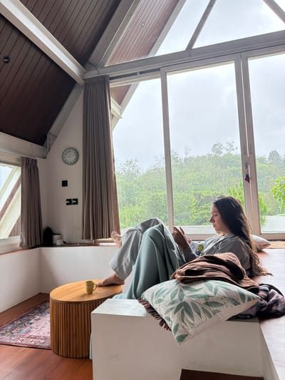 A woman sits on a window seat with pillows and blankets, facing forward and looking down at a device. Tall windows display lush greenery in the background. Inside, the room features wood-paneled walls, a clock, and shades. A round wooden table is next to the woman. The image is shot from an unusual angle, making the scene appear somewhat disoriented.