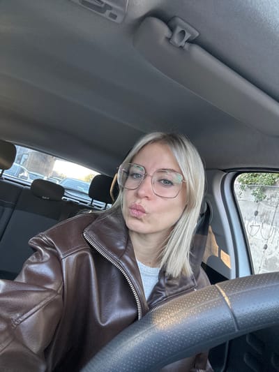 Inside a car, a young woman with blonde hair and glasses poses for a selfie. She wears a brown leather jacket over a white shirt. The car's interior, including the dashboard, steering wheel, and back seats, are visible. The woman has a slightly puckered expression. Outside the window, cars are parked in what appears to be a parking lot.