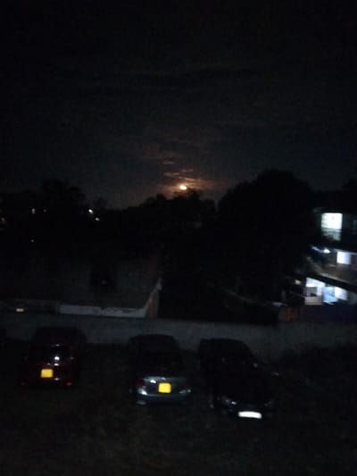This image depicts a nighttime scene in Riabai ward, Kenya. The overall environment is dark, illuminated primarily by a prominent, partially obscured full or near-full moon in the upper-middle sky, casting a warm yellow-orange glow through thin clouds. The sky above is dark grey to black with visible cloud formations.
In the foreground, a dark, unpaved or grassy area serves as a parking space for three dark-colored passenger vehicles. From left to right: a dark red or maroon sedan/hatchback with a visible yellow license plate at the rear; a dark grey or blue sedan with a visible yellow license plate at the rear; and a dark-colored sedan or SUV, partially visible on the right, facing towards the right with a faint light reflection on its front bumper. No individuals are visible in proximity to the vehicles.
The midground features a dark expanse of tree foliage and dark, indistinct terrain. A lighter-toned, horizontal structure, possibly a wall or pathway, is faintly discernible on the left side, separating the foreground from the dense foliage.
In the background, particularly on the right side, a multi-story building, likely residential or commercial, is visible. The building's structure is largely obscured by darkness, but multiple windows on its lower and upper floors are brightly illuminated with cool white light. Other distant, faint lights are scattered across the horizon, suggesting additional structures or streetlights amidst the silhouetted tree line. No human activity or interaction is discernible within the frame.