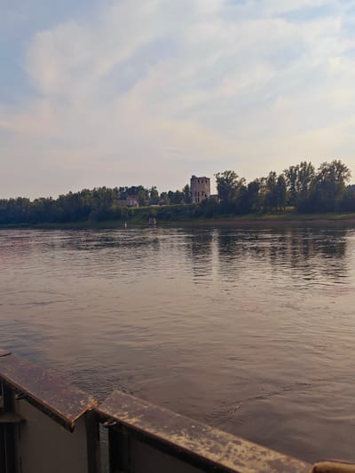 A body of water, likely a river, fills the foreground and middle ground of the image, reflecting the sky and the distant treeline. On the far bank, a dense line of green trees stretches horizontally. Further back, atop a gentle rise, are several structures. The most prominent is a tall, dilapidated stone building with a square base and visible damage, appearing to be an old tower or industrial remnant. To its left, other smaller buildings are visible, partially obscured by the foliage. The sky above is a pale blue with streaks of white clouds. In the lower left corner, a dark, rusted metal railing or barrier is visible, suggesting the vantage point is from a boat or a pier. The overall lighting suggests it is either early morning or late afternoon. The location is identified as the city of Yukseyevo, Russia.