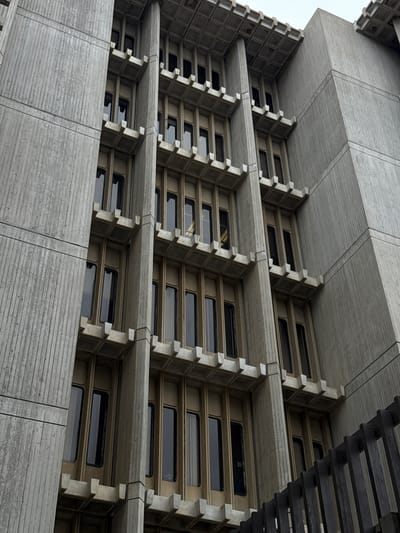 The image shows a low-angle, worm's-eye view of the exterior of the Brutalist architecture building known as the Salk Institute for Biological Studies, located in San Diego, California, United States of America. The building's facade is characterized by its strong vertical and horizontal lines, with repetitive concrete modules and deep-set windows. The concrete surfaces exhibit a textured, ribbed pattern, a common feature of Brutalist design. The windows appear dark and recessed, creating a sense of shadow and depth. The sky is overcast, suggesting a cloudy or diffused lighting condition. A dark, slatted fence or railing is partially visible in the lower right corner of the frame, in front of the building. There are no people visible in the image.