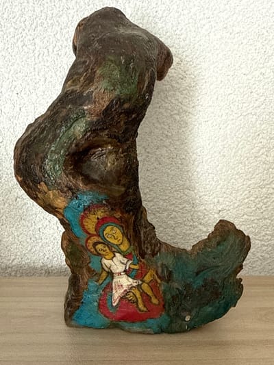 The image shows a piece of carved wood, likely a sculpture, with a painting of two figures on it. The figures appear to be religious in nature, possibly depicting the Virgin Mary and child, rendered in a style reminiscent of icons. The wood itself has a rough, textured surface and is carved into an abstract, somewhat anthropomorphic shape. The painting is colorful, with blues, reds, yellows, and skin tones used to depict the figures and their halos. The sculpture is placed on a wooden surface, with a textured white wall in the background. The lighting suggests an indoor environment. There are no discernible indications of location within the city of Belo pole, Bulgaria, or any other specific place.