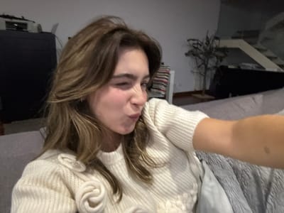 A young woman with long, light brown hair winks and puckers her lips in a selfie. She wears a cream-colored sweater with decorative floral details. The background includes a dark cabinet, a small tree-like plant, and a staircase. The photo is taken indoors, likely on a couch or sofa, suggested by the grey fabric. The lighting is soft, and the overall composition is casual.