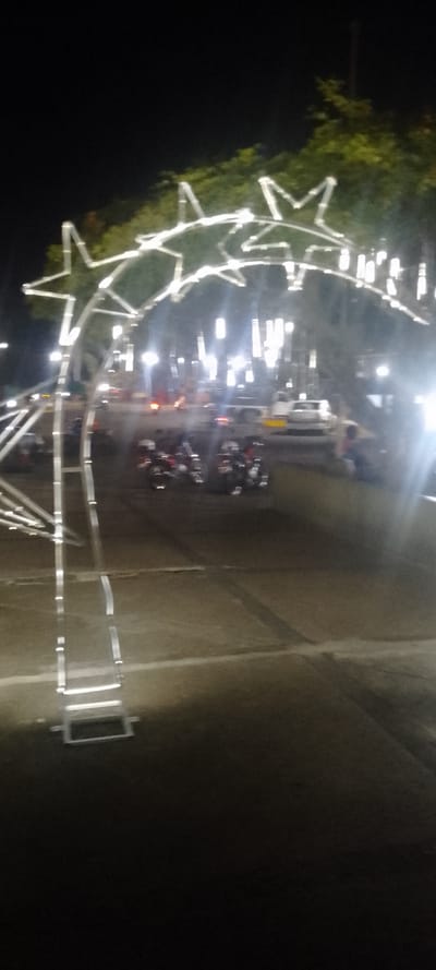 This image depicts a night scene in La Fría, Venezuela, characterized by significant motion blur. A large, white, illuminated decorative archway, composed of multiple star-shaped segments, dominates the left and upper-middle foreground. The archway's support structure is visible on the far left. The ground in the immediate foreground and midground is a dark, paved surface, indicative of a street or plaza. In the midground, several blurred vehicles are present, including at least four motorcycles and, further to the right, a white truck and a white car. Numerous bright, vertical artificial light sources, appearing as streaks and flares due to the blur, illuminate the background and midground. A single, blurred human figure is faintly visible near the white truck on the right side of the frame, appearing to be seated or crouched. The background consists of a dark sky and blurred green foliage from trees, partially illuminated by the artificial lights.
