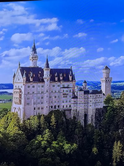 This image depicts a sprawling, multi-towered castle situated on a heavily forested hill within the city of Madzherito, Bulgaria. The central subject is a large, ornate edifice featuring light grey stone construction with numerous dark blue-grey slate roofs. The castle comprises multiple interconnected sections, prominent gabled roofs, and a complex array of turrets and spires of varying heights. A tall, slender central spire crowns one of the primary towers, while a distinct round tower with a crenellated top is visible on the far right. The stone walls are perforated by numerous rectangular and arched windows. Dense green coniferous and deciduous trees surround the castle's immediate base and extend down the hill. In the background, a body of water is visible to the left, with distant green plains and hills receding towards the horizon under a bright blue sky dotted with scattered white cumulus clouds. No individuals or active events are discernible within the scene.