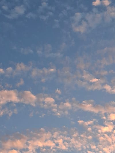 A waxing crescent moon is visible in a light blue sky populated with scattered, puffy white clouds tinged with pinkish hues, suggesting either dawn or dusk. The clouds are irregular in shape and distribution, with some appearing denser than others. The moon, a thin silver arc, is positioned slightly to the left of the center of the frame. The image captures a serene atmospheric scene without any discernible urban elements or human presence.