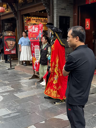 This photograph captures a daytime street scene in Qingyang District, China, featuring multiple individuals and commercial signage. In the foreground, a man stands facing left in profile, observing the scene. He wears a short-sleeved black polo shirt and black trousers with the word "CHINA" embroidered in red on the left thigh. He has short dark hair, spectacles, and light facial hair. His arms are crossed over his chest. Immediately to the left of this man, a performer in an elaborate traditional Chinese costume stands. The costume is predominantly red with extensive gold embroidery depicting dragons and intricate patterns. The performer wears a large black hat with gold accents, and their face is obscured by what appears to be a mask or heavy theatrical makeup. They wear black traditional shoes with gold embroidery. Behind and slightly to the right of the costumed performer is a woman in a light green traditional-style tunic or dress, covered by a black multi-pocketed sleeveless vest. She wears white athletic shoes and holds a red fan in her right hand. Her dark hair is pulled back. Further left, a large, rectangular red advertising sign is prominently displayed. The sign features the Chinese characters "川剧变脸" (Sichuan Opera Face-Changing) in a stylized font, with "18" in a red circle at the top right, possibly indicating a price or age. The sign lists various program elements such as "功夫茶艺" (Kung Fu Tea Ceremony), "变脸" (Face-Changing), "独家手影" (Exclusive Hand Shadows), "长绸水袖" (Long Sleeve Dance), "皮金" (Pijin opera/puppet show), and "古事" (ancient stories), alongside a cartoon illustration of a traditional character. To the left of the red sign, a woman in a light blue short-sleeved top and a white tiered skirt stands, observing the scene. She wears dark slip-on shoes. On the far left, a tall, vertical black poster with a red border displays a close-up image of a man's face, possibly a celebrity, wearing a red hoodie. Text on this poster includes "Jay" and "实体CD 限量版" (Physical CD Limited Edition). Partially visible behind this poster is a person wearing a dark jacket and blue jeans with white sneakers. The background consists of traditional-style buildings. The building on the left has a dark entrance and an overhead sign with a guitar icon and Chinese text that translates to "If music is a book, then CD is the beginning." A red lotus-shaped lantern hangs from the building's facade. The building on the