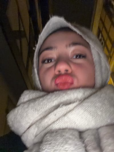 A person with fair skin, dark eyes, and dark eyebrows is centered in the frame, wearing a light gray, hooded scarf wrapped around their head and shoulders. They are pouting their lips in a playful manner. The camera angle is slightly tilted. The background appears to be dimly lit, suggesting an indoor or nighttime setting.