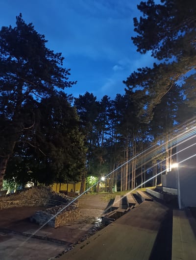 The image depicts an outdoor scene at dusk or night, with a clear, deep blue sky visible above a dense line of tall pine trees. In the foreground, there is a stone structure that appears to be ancient ruins, possibly with curved walls made of rough stones. To the right, a modern wooden staircase with several steps ascends, leading to a building with a grey facade illuminated by bright lights that cause lens flare. Scattered picnic tables and benches are visible in the background amongst the trees. The ground is a mix of dirt, gravel, and possibly paved areas. The overall atmosphere suggests a public park or an archaeological site within a wooded area, possibly near an urban center, given the presence of the building and artificial lighting. Based on the visual cues and the location provided, this scene likely represents the Archaeological Complex "Constantine and Helena" in Kyustendil, Bulgaria, which features ancient Thracian and Roman ruins situated amidst pine trees and often includes modern amenities for visitors.