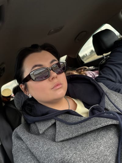 A woman with dark hair and sunglasses is sitting in the passenger seat of a car. The car has a sunroof. The woman is wearing a grey coat over a dark blue hooded sweatshirt and a cream-colored shirt. She is also wearing a silver chain necklace and small, light-colored earrings. The environment visible through the sunroof and car window indicates an outdoor setting. Based on the prompt, the location is Yugo-Kamskiy, Russia.