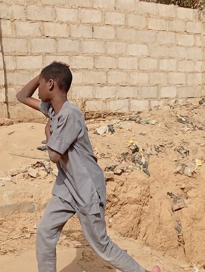The scene depicts a young male individual in Fagge D2, Nigeria, captured mid-stride or movement. The subject, an adolescent with short dark hair, is dressed in a light grey two-piece traditional garment consisting of a short-sleeved tunic and matching trousers. His right hand is raised to the back of his head, while his left arm is bent at the elbow and held across his body. He is positioned facing left within the frame, with his left leg extended forward and his right leg bent, suggesting motion. The background features a light-colored concrete block wall, partially visible. The ground is uneven, composed of sandy earth, with a visible mound of debris to the right containing scattered plastic bags (black and yellow) and other refuse. Sparse dry vegetation is visible at the base of the wall and among the debris. Bright daylight illuminates the scene, casting a shadow from the individual onto the ground.