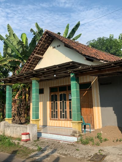 The image depicts the front of a single-story house on a bright, sunny day, likely in the city of Tanjungsepreh, Indonesia, given the tropical vegetation. The house has a light yellow exterior with vertical cream-colored stripes. A covered porch extends across the front, supported by two square pillars that are decorated with green tiles and topped with yellow bases. The roof of the porch is made of corrugated metal. The main roof of the house is tiled with terracotta tiles, and a wooden overhang is visible.

In front of the house, there is a paved walkway and a low concrete wall. To the left of the walkway, a small, red-and-white decorative element sits on the ground. Lush green banana trees and other tropical foliage are prominent behind and to the left of the house. The house features a wooden front door and a large window with multiple panes. A teal plastic chair is placed near the entrance. A pile of dirt is visible to the right of the house, suggesting recent construction or landscaping. The ground is a mixture of paved stones and patches of grass. The sky is blue with scattered white clouds. There are no people visible in the image.