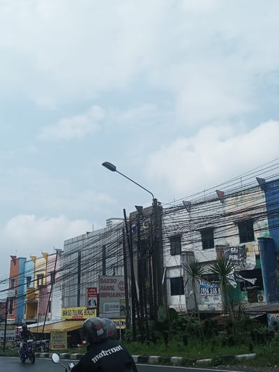 A street scene in a bustling Asian town shows rows of colorful buildings adorned with tangled power lines. Advertisements for "Bakso Tulang Bogor" and other local businesses are visible. A lone street lamp stands tall amidst the electrical chaos. Two motorcyclists are riding on the road in the foreground, one wearing a jacket with "maternal" printed on the back. Lush greenery peeks through along the roadside. Above, the sky is a pale blue with scattered clouds.