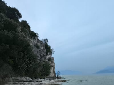 The image depicts a coastal scene in Sirmione, Italy, characterized by a sheer, layered rock cliff rising from a pebbled shore. Lush green vegetation, including trees and bushes, clings to the upper portions of the cliff face and spills down towards the water. The sea is calm and stretches towards a hazy horizon where distant mountains are visible. The sky is overcast and a muted blue, contributing to the serene atmosphere. On the shore, a single individual is standing near the water's edge, facing away from the camera. The overall impression is one of natural beauty and tranquility.