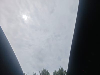 The image depicts a cloudy sky with the sun partially obscured by clouds, creating a bright, diffused light source. Two dark, angular structures frame the sky from the left and right sides of the image, creating a V-shape. The lower portion of the image shows the tops of green trees, suggesting an outdoor environment. There are no people or specific objects of interest visible in the sky or among the trees. The scene appears to be captured from a ground-level perspective, looking upwards. The lighting conditions are overcast, with the sun providing a soft illumination. The overall impression is one of a natural outdoor setting under a cloudy sky, possibly in a park or wooded area within the city of Lyublinskiy Sadovod, Russia.
