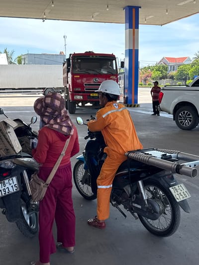 This media file captures a scene at a gas station in Hòa Đa, Vietnam. Two individuals are the primary subjects. The first is a person wearing an orange jumpsuit and a white helmet, appearing to be working at the station. They are leaning over a motorcycle. The second individual is wearing a red top, maroon pants, and a patterned headscarf, also near a motorcycle. The setting includes a red truck, a white van, and the characteristic blue and orange striped structure of a gas pump.

The time of day appears to be daytime, with bright sunlight casting shadows, suggesting a clear or partly cloudy sky. The atmosphere seems to be that of everyday activity at a service station, with no overt display of strong emotions.

Visible text includes license plates on the motorcycles and a truck. One motorcycle's plate reads "78-C1 193.15" and "78-C1 475.61". The truck's plate is "78C-077". The word "HONDA" is visible on the motorcycles, and "YUNSITE" is present on a strap worn by the second individual. The gas station structure itself has blue and orange bands.