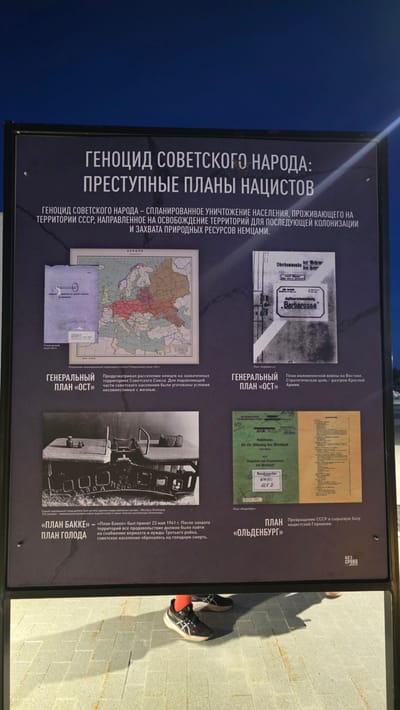 Here's a detailed description of the image:
The image shows a display board, likely part of an exhibition, presenting information about the Nazi plans for the genocide of the Soviet people. The board is divided into sections, each containing text in Russian and related images. The main title reads: "Genocide of the Soviet People: Criminal Plans of the Nazis."
The accompanying text elaborates on the planned destruction of the population living in the territory of the USSR, aimed at freeing up territories for subsequent colonization and the seizure of natural resources by the Germans.
Specific plans mentioned and illustrated on the display board include:
* **"General Plan Ost":** This section features a map of Eastern Europe with different colored areas, presumably indicating regions designated for German resettlement and Soviet population displacement. There is also a picture of a document with text on it. The accompanying text says the plan envisioned the resettlement of Germans in the captured territories of the Soviet Union, with the conditions for the majority of the Soviet population being incompatible with life. It is described as a plan for a lightning war in the East, with the strategic goal of defeating the Red Army.
* **"Plan Oldenburg":** Another section features a document and text that states the plan was for turning the USSR into a raw material base for Nazi Germany.
* **"Bakke Plan" ("Hunger Plan"):** This part of the display board includes a photograph of railway cars and describes the "Bakke Plan" as a plan to supply the Wehrmacht (German army) and the needs of the Third Reich, with the consequence that the Soviet population was doomed to starvation. This plan was adopted on May 23, 1941.
In the reflection of the display are legs of a person wearing white/black shoes and a bright orange sock. Based on the information provided, it is not possible to determine the exact location in Zaitsevo (Verevskoe S/P), Russia, but it is likely inside a museum or memorial complex that focuses on WWII history.