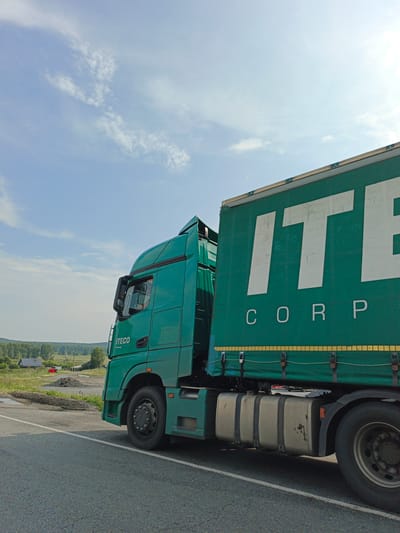 A green semi-truck is parked on the side of a road. The truck has a large green trailer with the white letters "ITECO CORP" printed on the side. The truck cab is also green and has "ITECO" printed on the door. The truck is facing to the right of the frame, and the trailer is extending out of the right side of the frame. In the background, there is a field with some trees and a few buildings. The sky is blue with some white clouds. The location is Nizhnii Atlyan, Russia.