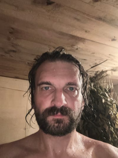 The media file is a close-up, selfie-style shot of a man with wet, dark hair and a beard, looking directly at the camera. His eyes are a striking blue-green, and his face is slightly flushed, with visible sweat droplets on his skin. He appears to be in a sauna, indicated by the wooden paneling of the walls and a bundle of leafy branches, likely a venik, resting above and behind him. The lighting is warm and somewhat dim, suggesting an indoor setting. The man's expression is relaxed, perhaps a bit contemplative or invigorated. The background is out of focus, but the wooden textures are prominent. There is no visible text in the image. The overall impression is one of relaxation and well-being in a traditional sauna environment.