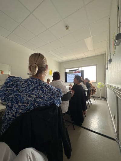 This image depicts an indoor scene in a meeting or conference room located in Tourcoing, France. The room features light-colored walls, a tiled drop ceiling with a smoke detector, and a light grey floor. A large window at the far end illuminates the space.
Seven individuals are visible. In the immediate foreground, a person with blonde hair in a ponytail, wearing a blue and white floral top, is seated with their back to the viewer. A black jacket is draped over their chair. To the left, a light-colored utility area with white tiled walls is partially visible.
In the midground, several people are seated around a dark table. One individual, wearing a white uniform or shirt, is seated to the left of the table. To the right, a person with dark hair, wearing a black top, is actively observing a projection screen. The screen displays a presentation slide titled "VISA : conclusion" with text below. Further individuals are partially visible around the table, engaged in the activity or conversation.
A light-yellow standing fan is positioned on the floor near the right side of the projection screen. Along the right wall, a horizontal metal railing or guard bar runs at waist height, and a dark cable or tube extends along the floor. The overall scene suggests a group attending a presentation or meeting.