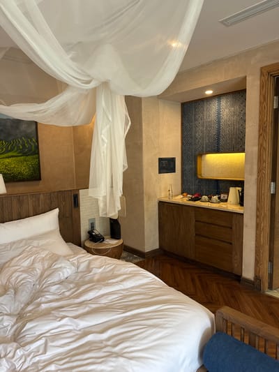 An interior view of a hotel room in Púng Luông, Vietnam, showcases a blend of natural textures and modern amenities. In the foreground, a large bed is made with white linens and pillows, its duvet appearing rumpled. Above the bed, a sheer white canopy or mosquito net is gracefully draped from the ceiling. To the left of the bed, a wooden headboard is visible, above which hangs a framed painting depicting vibrant green and yellow terraced fields. A small, round wooden nightstand to the right of the bed holds a black corded telephone, a black cylindrical object, and a white piece of paper. The walls surrounding the bed area are textured in an earthy brown-tan hue.
To the right, a dedicated mini-bar or coffee station is built into the wall. It features light-colored stone countertops over dark wooden cabinets with drawers and doors. The back wall of this station is adorned with a dark blue patterned wallpaper or fabric, displaying geometric designs. Mounted above the counter is a horizontal, rectangular light fixture emitting a warm yellow glow. On the countertop, various items are arranged: a glass water bottle, multiple pieces of fruit (including dragon fruit and a small orange), two white cups and saucers, a black tray, and a white electric kettle with a coffeemaker. A small black information plaque is affixed to the wall to the left of this station. The floor throughout the visible area is dark wood, laid in a herringbone pattern. A wooden door frame is partially visible on the far right, suggesting an adjacent room or entrance. The ceiling is white, with an air vent visible towards the upper right.