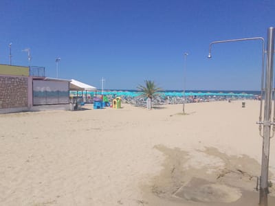 This is a wide, horizontal, outdoor shot of a beach on a clear, sunny day. The sky is a vibrant blue, and the sand is a light beige. In the foreground, a beach shower made of metal stands to the right. The sand around its base is wet and cracked, with some sparse green vegetation growing nearby.
Further back, a row of lounge chairs and umbrellas stretch across the beach, suggesting a popular tourist destination. The umbrellas are predominantly turquoise, with a few other colors interspersed. A small palm tree stands among the umbrellas. To the left, a building with a stone wall and a light yellow facade is visible, with a white awning extending from it. A metal fence with a balcony separates the building from the beach. Several colorful children's playground structures, including a blue and yellow slide and a red and green climbing structure, are located near the building.
In the distance, the calm sea meets the horizon under the clear blue sky. A few distant figures can be seen on the beach, but they are too small to discern details. There is no visible text in the image. The scene evokes a feeling of a warm, leisurely summer day at a well-equipped beach, likely in a Mediterranean location like Valle Castellana, Italy, given the context. The presence of the shower, chairs, umbrellas, and playground indicates a public beach facility.