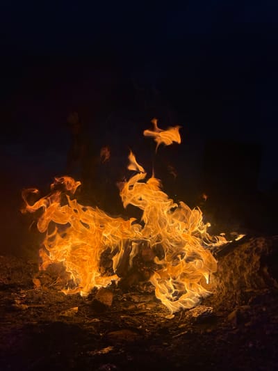 A close-up, low-angle shot captures a campfire burning intensely at night in Kaspiysk, Russia. The flames, predominantly shades of bright orange and yellow, are dynamically shaped by the wind, creating swirling patterns and licking upwards. Wisps of smoke are visible against the dark, inky sky, which takes up the upper portion of the frame. The ground beneath the fire consists of dark earth, scattered rocks, and charred debris. In the background, barely discernible shapes suggest the presence of distant structures or figures, softened by the darkness and the glow of the fire. The overall impression is one of raw, untamed energy emanating from the central blaze.
