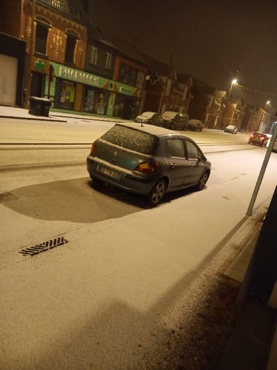 A car parks on a snow-covered street at night. The vehicle is angled towards the curb. To the left, illuminated shop signs and buildings line the street. Streetlights cast a warm glow on the surroundings. Snow covers the road and sidewalks. A storm drain is visible in the foreground. The scene suggests a quiet, snowy night in a residential or commercial area.