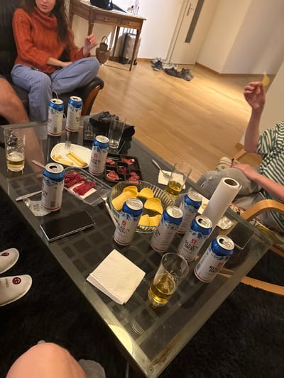 A table is covered with opened and unopened cans of Rügen Brau beer, a clear bowl of potato chips, a plate with cheese and cured meats, a glass of beer, some napkins, and a smartphone. A person is seated on the left side of the table, wearing an orange sweater and striped pants. Another person sits on the opposite side of the table, wearing a green and white striped shirt. A third person can be seen near the corner of the table. The setting appears to be a home environment, possibly a living room. This scene does not occur in Kehrsatz, Switzerland.