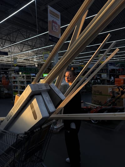 A young woman with her hair in a bun stands in the aisle of a hardware store in Yudinovo, Russia. She is wearing a black jacket and dark pants. She is positioned behind a shopping cart filled with long wooden planks and boxes. The lighting in the store is low, with bright fluorescent lights visible on the ceiling. In the background, shelves stocked with various items and other shoppers are visible. The scene is captured in a low-angle, wide shot.