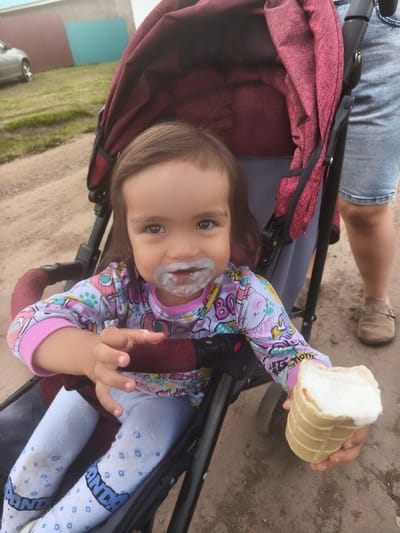 A young child, with white residue around their mouth, sits in a stroller holding an ice cream cone. The child is wearing a colorful long-sleeved shirt and light blue leggings with a pattern. A person's legs, wearing denim shorts and brown shoes, are visible to the right of the stroller. The scene appears to be outdoors, with a dirt path and a grassy area in the background. A silver car is parked on the left side of the frame, and a fence with blue and burgundy panels is visible behind it. The context is the village of Burayevo, Russia.