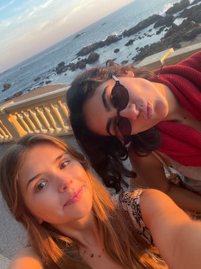 The image depicts two young adult females posing for a photograph on a coastal promenade in Aldoar, Foz do Douro e Nevogilde, Portugal, during late afternoon "golden hour" lighting.
The subject in the foreground on the left has long, light brown hair, is wearing a patterned top, and a subtle necklace. She looks directly at the camera with a slight smile. The subject positioned behind and to her right has dark, wavy hair, wears dark-lensed round sunglasses, and puckers her lips. She is draped in a red fabric over her shoulders.
The background displays the Atlantic Ocean with numerous dark, rugged rock formations breaking the surface, extending toward the horizon. A light-colored balustrade, illuminated by the warm, low-angle sunlight, separates the promenade from the ocean. The sky above is clear, transitioning from light blue to warmer tones near the horizon. The immediate foreground is a paved surface.