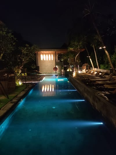 This image depicts a nighttime scene at a resort property in Mas, Indonesia. The central feature is a long, rectangular swimming pool, illuminated from within by blue underwater lights creating distinct beams. To the left of the main pool, a grassy area features tropical foliage and trees, with a decorative, arched, woven structure. A pathway, marked by ground-level lighting, runs alongside this section of the pool.
In the background, a multi-story building with modern and traditional architectural elements is illuminated. Its upper facade displays five vertically oriented, rectangular panels or windows glowing with warm light, which reflect distinctly in the pool's surface. Below this building, two individuals are partially visible in a connected, shallower pool or hot tub area.
To the right of the main pool, a raised wooden deck is visible, furnished with approximately twelve unoccupied light-colored lounge chairs arranged in a row. Several tall palm trees line this deck, some illuminated by upward-facing lights mounted on their trunks. Another decorative, woven circular chair or daybed is present on the deck further from the foreground. The overall environment is lush with tropical vegetation, silhouetted against the dark night sky.