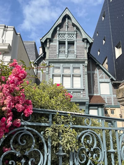 The image shows a detail of an ornate, historic building in Trouville-sur-Mer, France, viewed through a decorative wrought-iron fence and lush pink roses. The building's facade features intricate woodwork and shingle siding, painted in muted tones of grey-blue and green. A prominent gabled roof with decorative finials caps the structure. Adjacent to it, another building displays a dark, diamond-patterned slate roof and a white-framed window. The foreground is dominated by the vibrant pink blossoms of a rose bush and variegated green foliage, partially obscuring the ornate blue fence. The scene is bathed in bright daylight under a clear blue sky with scattered white clouds, suggesting a pleasant day in the coastal town.