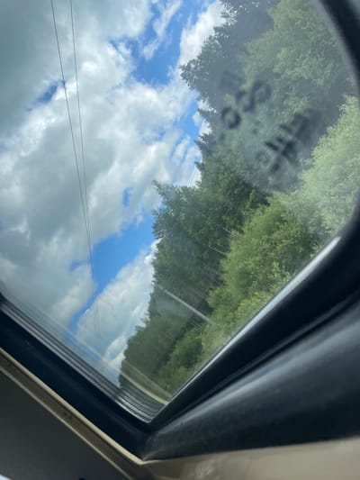 The image shows a view through a sunroof, looking up at a bright blue sky with scattered white clouds. Two thin, dark power lines stretch across the sky diagonally from the upper left to the lower right. To the right of the power lines, a dense forest of green trees fills the scene. Some of the trees are tall and slender, while others are bushier and shorter. The trees appear to be on a slope, with some further into the background seeming to be higher up. The bottom of the image shows the black frame of the sunroof and the light-colored interior of a vehicle. There is some smudged writing on the glass of the sunroof, partially obscuring the view of the trees. The scene is likely captured while the vehicle is in motion, possibly on a road surrounded by forest. The location is identified as Elovo, Russia.