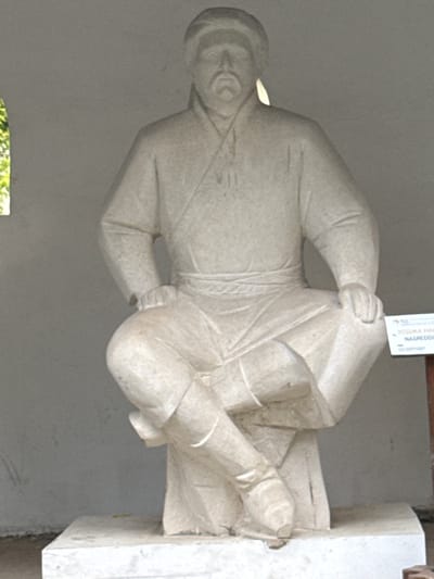 The image shows a stone statue of a seated man wearing traditional attire, including a cap, tunic, and boots. The statue is light in color and appears to be weathered. The man has a mustache and is looking forward. He is seated on a pedestal. To the right of the statue, a small white sign with text in Cyrillic script is visible. The background is a plain gray wall, suggesting an outdoor or semi-outdoor location. A small section of greenery is visible on the left side of the image, peeking through an opening in the wall. The statue is positioned in the city of Petrovo, Russia, and appears to be a historical monument.