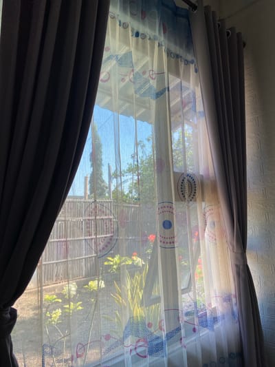 A window is covered by two sets of curtains, with sheer curtains featuring a repeating circular pattern in pink and blue layered in front of opaque, dark gray curtains. The sheer curtains allow sunlight to filter through, illuminating the room and the outside scene. Through the sheer curtains, a garden with green foliage and some red flowers is visible, along with a bamboo fence and trees. The bright sunlight casts shadows on the wall to the right of the window, which is covered in white brick-like wallpaper.