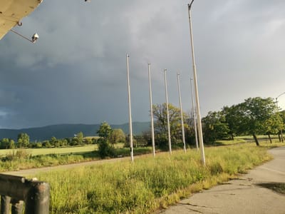 The image captures a rural landscape in Zhivkovo, Bulgaria, under a dramatic sky. The scene is dominated by a clear distinction between a dark, stormy sky on the left, hinting at recent or impending rain, and a brighter, lighter grey sky on the right, where sunlight appears to break through. This contrast suggests late afternoon or early evening.
In the middle ground, a series of seven tall, slender white poles stand vertically, some with small black attachments or fittings. These poles divide the frame and cast long, faint shadows to the right. Beyond the poles, lush green fields stretch towards a range of dark, undulating mountains that form the backdrop. Scattered trees with varying shades of green foliage are visible across the middle ground and right side of the image.
In the foreground, the ground is covered in dense, overgrown green and yellow-green grass, bordering an asphalt road or path that curves from the right foreground into the middle ground. A dark, possibly rusty, metal structure, perhaps part of a railing or gate, is partially visible in the immediate lower left corner. In the upper left, a light-colored building overhang with an attached security camera or light fixture is visible, with another small light fixture or sensor appearing against the sky in the upper right.
No people or discernible activities are taking place, presenting a tranquil yet atmospheric view of the natural and semi-rural environment. There is no visible text in the image.