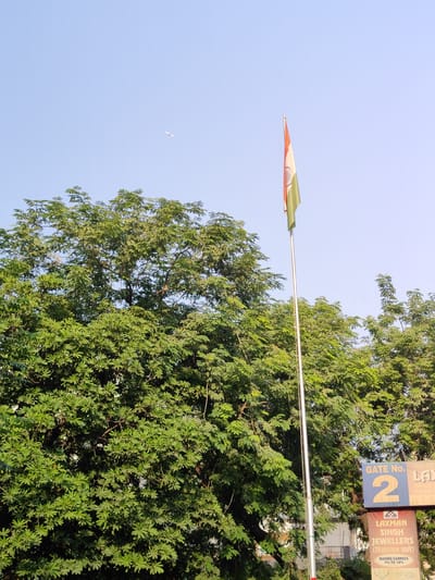 The image shows an Indian flag flying high on a flagpole against a clear blue sky, with lush green trees in the foreground. The flag is partially unfurled and displays the saffron, white, and green bands with the Ashoka Chakra in the center. In the far distance, a small airplane can be seen in the sky. At the bottom right of the image, there are signs indicating "GATE No. 2" and "LAXMAN SINGH JEWELLERS (हिरा और वाले)". The scene appears to be an outdoor environment in Gurgaon, India, likely near a commercial area or entrance.