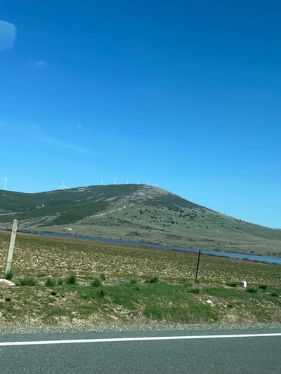 The image depicts a wide landscape view in Bernuy-Salinero, Spain, under a clear blue sky. In the foreground, the bottom edge features an asphalt road with a solid white painted line. Immediately above the road is a grassy verge with patches of dirt and sparse green vegetation. Two vertical posts are visible within this verge: a rectangular, grey concrete post on the left and a thinner, dark brown or black metal post further right.
The midground is occupied by a broad, relatively flat field exhibiting a mix of dry, light brown soil and scattered patches of green grass. A significant body of water, appearing dark blue, extends horizontally across the scene, separating the field from a distant hillside. A small, light-colored building with a distinct reddish roof is discernible on the far bank of this water body, nestled at the base of the hill.
The background is dominated by a large hill or mountain range. Its slopes are covered with a mix of dark green trees and lighter green scrub vegetation. Along the crest of this hill, approximately eleven white wind turbines are arrayed, their blades visible against the sky, some appearing to be in various stages of rotation. The upper two-thirds of the image consist of a uniform, clear blue sky with no visible clouds. No individuals or active subjects are present in the scene.