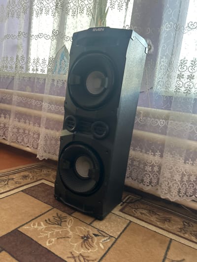 The image shows a large, black, rectangular speaker system with two visible circular speaker grilles. The speaker is positioned horizontally. On the left side of the speaker, partially obscured, is the word "SVEN" in white text. The speaker is placed in what appears to be a living room setting. Behind the speaker, white lace curtains with a floral pattern are visible, covering a window or glass door. The curtains are semi-transparent, allowing a soft, diffused light to enter the room, suggesting it is daytime. The floor is covered by a patterned carpet in shades of beige and brown. The overall scene is domestic and quiet. There are no people or discernible activity taking place in the image. The lighting is soft and even. The location cues, such as the carpet pattern and curtain style, are consistent with an indoor domestic environment.